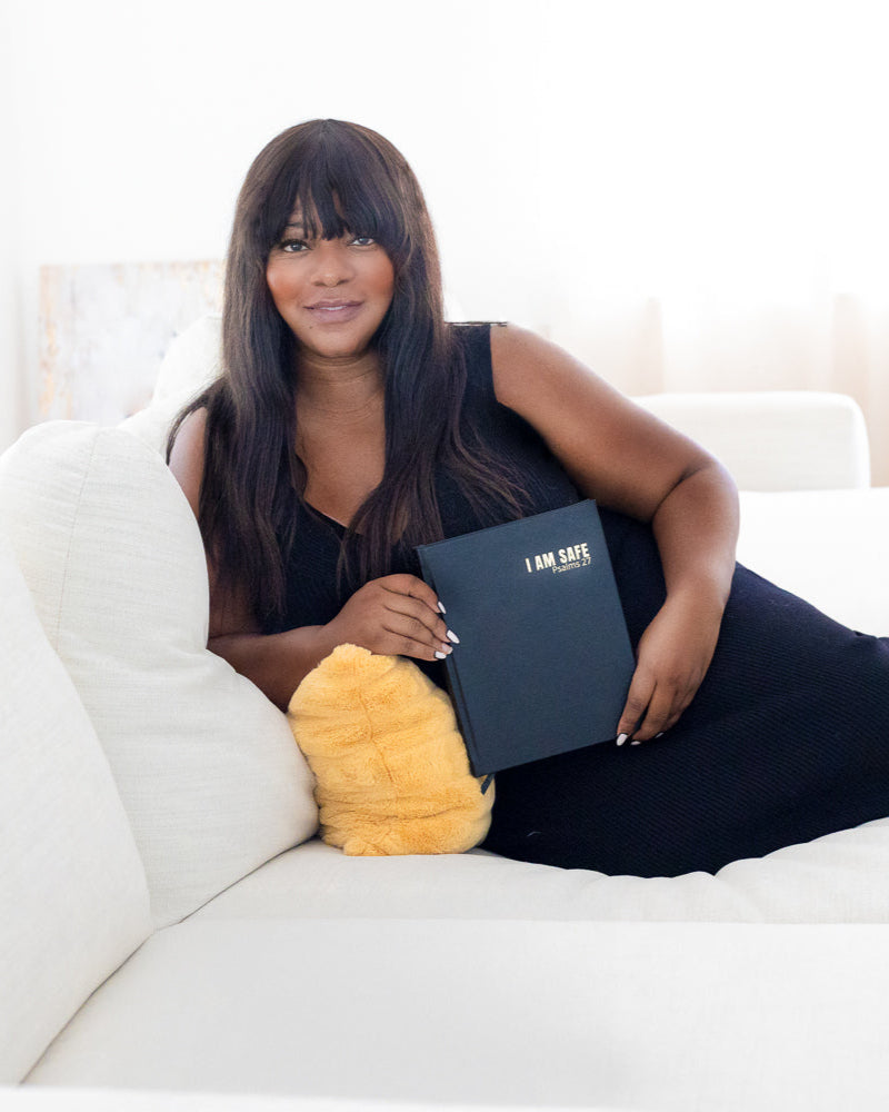 Woman sitting on a white couch holding a book journal " I AM SAFE" with a yellow pillow next to her.