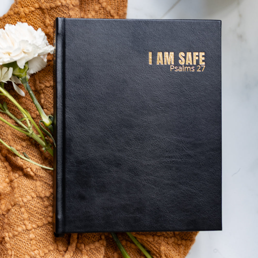 Black notebook with 'I AM SAFE Psalms 27' text on a textured surface with flowers and a mug.
