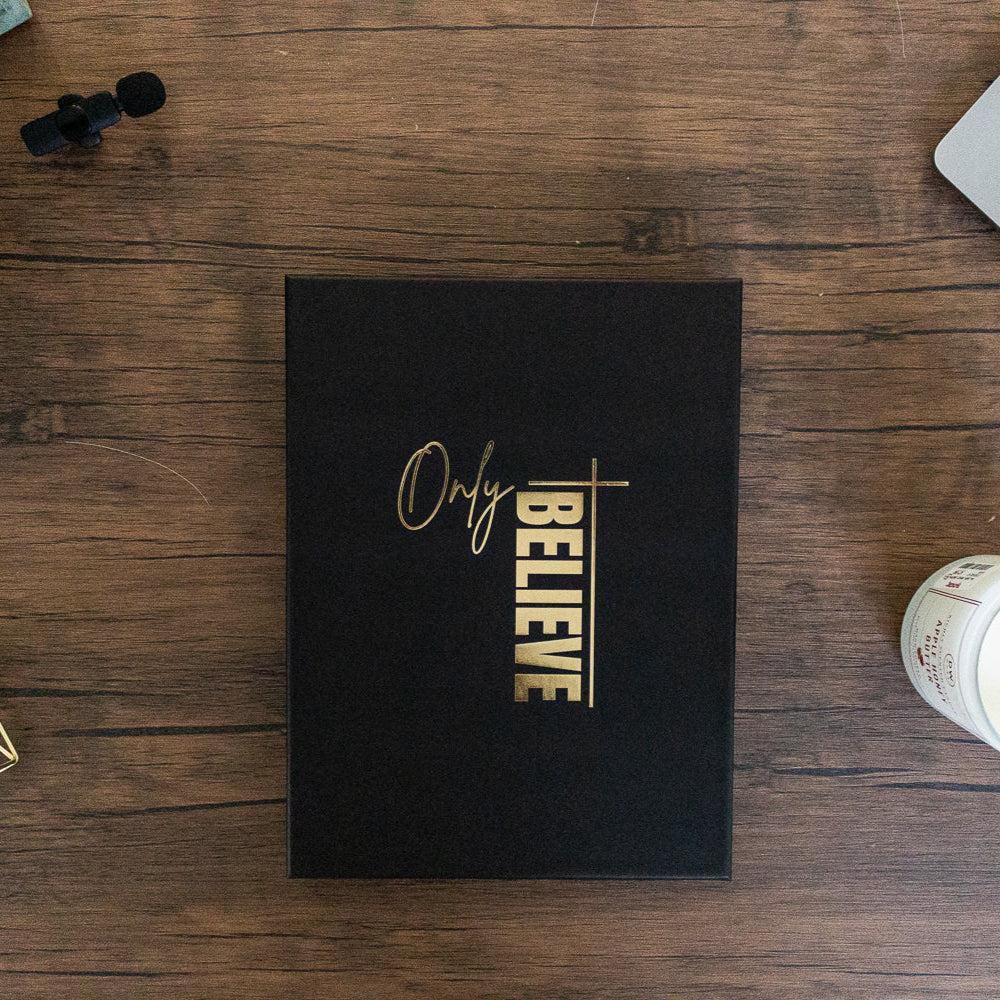 Black book with 'Only Believe' on a wooden desk with a laptop and decor items.