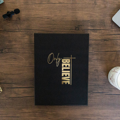 Black book with 'Only Believe' on a wooden desk with a laptop and decor items.